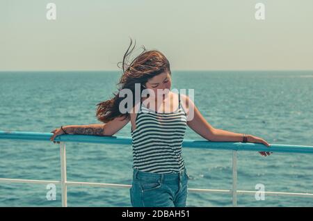 pretty young brunette girl with fluttering hair in a striped t-shirt and blue denim shorts enjoys the sea on the deck of a ship Stock Photo