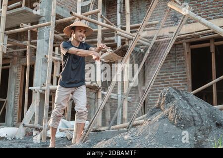 mason holding shovel while carrying sand to sieve against the backdrop of house building Stock Photo