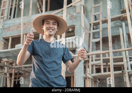 The mason wears a smiling cap with a thumbs up and holds a cement taper to the workplace Stock Photo