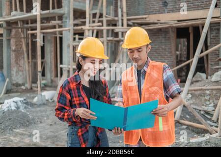 male and female contractors standing holding sketchbooks of drawings wearing safety helmets against the background of the unfinished building Stock Photo
