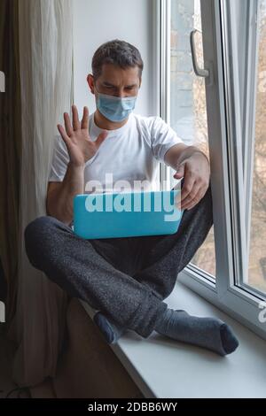 Stay Home work place. Man using laptop, working or chatting from home during quarantine sitting on the windowsill near the window. Stay home stay safe Stock Photo
