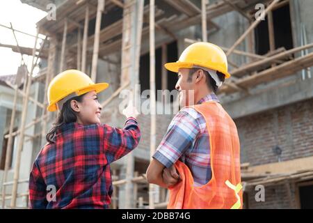Asian male and female contractors discussing while pointing fingers wearing safety helmets with the background of the unfinished building Stock Photo