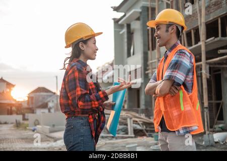 two Asian contractors discussing with hand gestures wearing safety helmets and vests against the background of an unfinished building Stock Photo