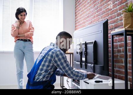 Young Woman Sitting On Couch Looking At Male Technician Repairing TV At Home Stock Photo