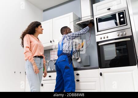 Male Repairman Checking Extractor With Digital Multimeter While Woman Standing In Kitchen Stock Photo