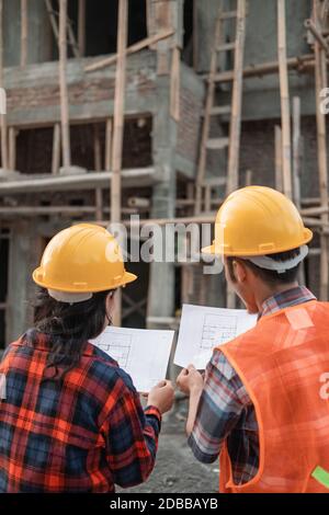 view from behind of male and female asian contractors standing wearing safety helmets holding the site plan building in the background of the unfinished building Stock Photo