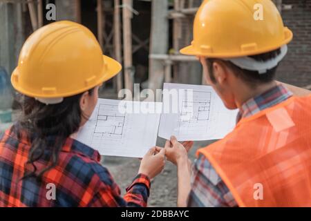 view from behind of two Asian contractors standing in safety helmets holding the site plan building in the background of the unfinished building Stock Photo