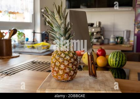 A cutting board with a whole pineapple fruit and a cleaver standing on a worktop in the kitchen in the background are more fruits and kitchen utensils Stock Photo