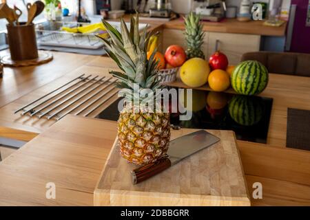 A cutting board with a whole pineapple fruit and a cleaver standing on a worktop in the kitchen in the background are more fruits and kitchen utensils Stock Photo
