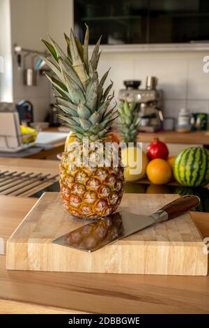 A cutting board with a whole pineapple fruit and a cleaver standing on a worktop in the kitchen in the background are more fruits and kitchen utensils Stock Photo