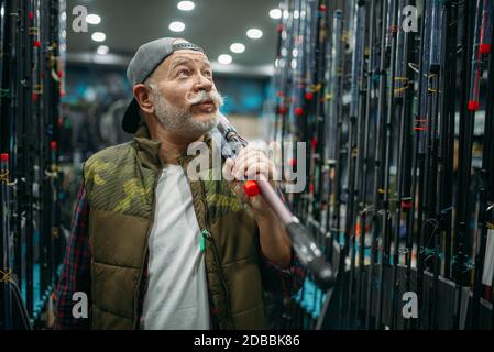 Male angler holds rod in fishing shop. Equipment and tools for fish catching and hunting, accessory choice on showcase in store, spinnings and telesco Stock Photo