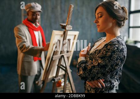 Portrait painter at the easel draws female model. Male artist standing at his workplace, creative master works in workshop Stock Photo