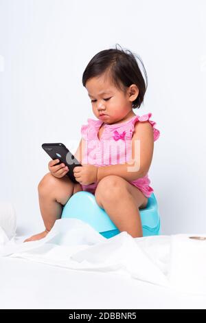 Asian little cute baby child girl education training to sitting on blue chamber pot or potty and play smart mobile phone with toilet paper rolls, stud Stock Photo
