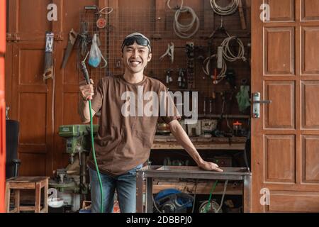 smiling welder looks into the camera while holding an electric welder in a welding shop Stock Photo