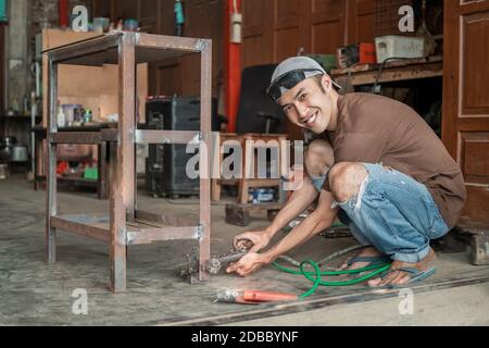 male welder smiles while holding the negative pole clamp for the electric welder on the metal frame Stock Photo