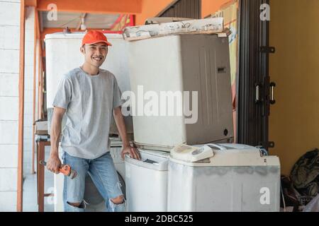 Asian electronics worker smiles at the camera holding a wrench standing next to a broken washing machine at an electronics service shop Stock Photo