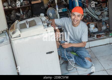 Asian male electronics worker smiles at the camera while repairing a broken washing machine cord at an electronics repair shop Stock Photo