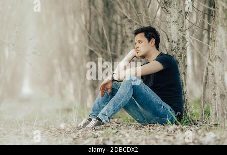young man sitting by a tree in the woods thinking about life and resting Stock Photo