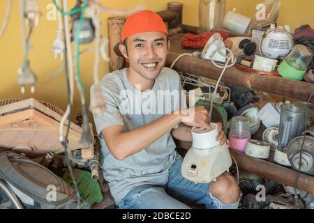 electronics repairman smiles at the camera while holding a broken blender against the backdrop of a shelf of broken goods in a service shop Stock Photo
