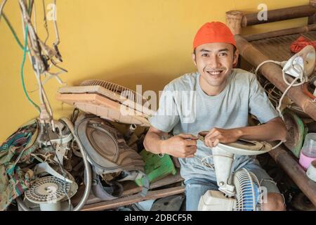 electronics repairman smiles at the camera while holding the fan cable while sitting around broken items in a service shop Stock Photo