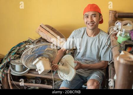 Asian electronics repairman smiles at the camera while holding the fan while sitting around broken items in a service shop Stock Photo