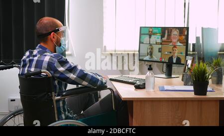 Disabled expert financial in wheelchair talking with partners wearing mask and visor during video conference in business office. Immobilized freelancer working in company respecting social distance Stock Photo