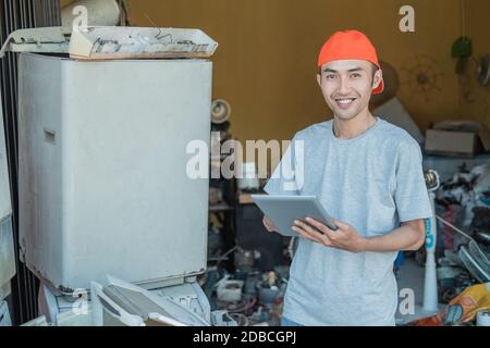 asian electronics repairman smiles at the camera while using his tablet pc around broken electronics in service shop Stock Photo