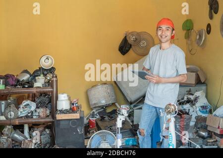 asian electronics repairman smiles at the camera while using his tablet pc around electronic parts in the service shop room Stock Photo