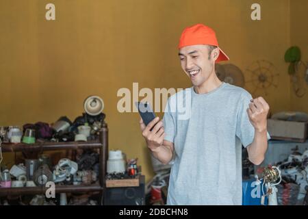 Asian electronics repairman with an excited gesture when looks at the smartphone screen while standing around the electronic parts Stock Photo