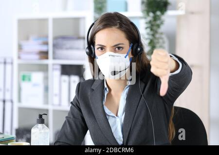 Angry telemarketer woman with thumbs down avoiding covid-19 looking camera with mask sitting in the office Stock Photo