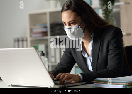Concentrated executive woman working late on laptop avoiding covid-19 with mask sitting at night in the office Stock Photo
