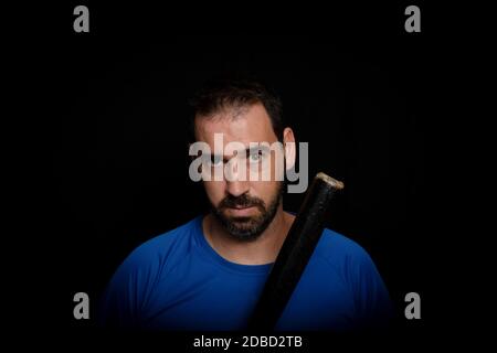 Bearded man dressed in blue t-shirt in defiant pose posing with a baseball bat on a black background.Threat concept Stock Photo