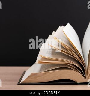 A large, thick hardcover book lies on a light-wood table. The book is open, the leaves of the page are fanned out, and the back is a black background Stock Photo