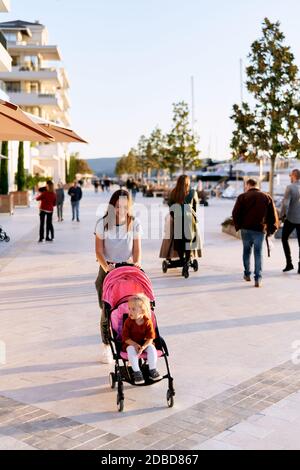 Woman is pushing a pink stroller with her daughter sitting inside of it while having a walk on a boat pier in Montenegro Stock Photo