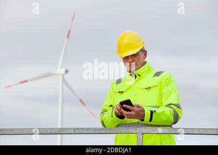 Mature engineer with protective clothing looking at a smart phone in front of a wind turbine in the blurred background Stock Photo