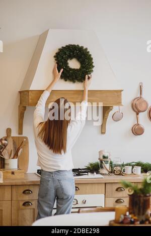 Back view of beautiful woman in white sweater hanging a Christmas wreath on her kitchen. Stock Photo