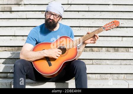 The funny bearded man with grimace is playing guitar and sitting on stairs. Stock Photo