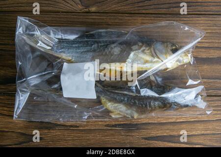 Dried salted crucian fishes in a transparent package with a white label on wooden background. Snack to beer. Stock Photo