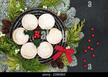 Festive homemade Christmas mince pies on a plate with holly, snow covered fir, mistletoe, cedar cypress, bow & loose berries on grey grunge background Stock Photo