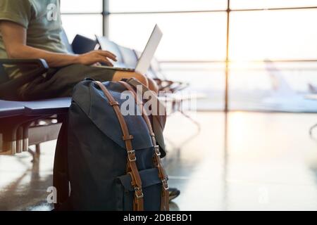 asian man male air traveler sitting in waiting area in airport terminal building using laptop computer, focus on the backpack Stock Photo