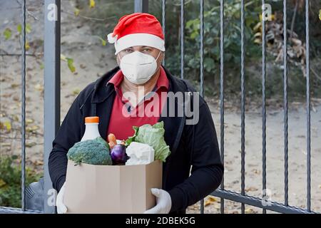 https://l450v.alamy.com/450v/2dbek5g/delivery-during-quarantine-o-food-donation-concept-man-in-santa-claus-hat-protective-mask-and-medical-gloves-holding-cardboard-box-with-various-food-2dbek5g.jpg