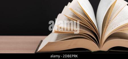 A large, thick hardcover book lies on a light-wood table. The book is open, the leaves of the page are fanned out, and the back is a black background Stock Photo