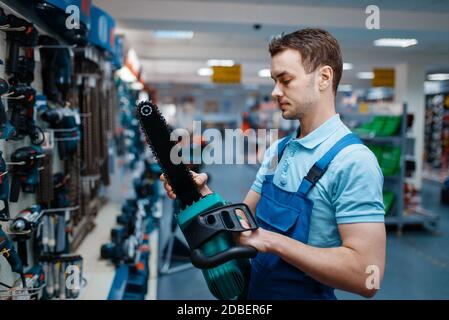 Male worker in uniform holds electric saw in tool store. Choice of professional equipment in hardware shop, instrument supermarket Stock Photo