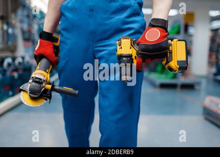 Male worker holds battery powered screwdriver and angle grinder in tool store. Choice of professional equipment in hardware shop, electrical instrumen Stock Photo