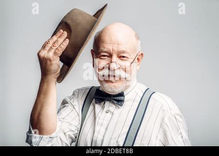 Elderly man in a bow tie and glasses takes off his hat, grey background. Mature senior looking at camera in studio Stock Photo