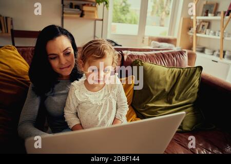Young mother and kid daughter having fun shopping online using laptop sitting on sofa