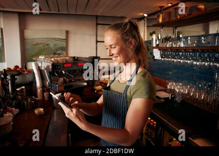 Young female staff wearing apron working with a digital tablet at the counter of cafe Stock Photo