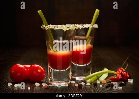 Two shot glasses with cocktail Bloody Mary, celery, spices and ripe tomatoes on dark wooden background Stock Photo