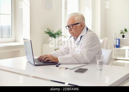 Serious white-haired general practitioner working on laptop sitting at desk in hospital office Stock Photo