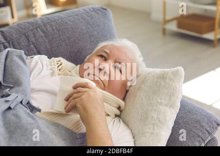 Sick senior man with a scarf wrapped around his neck lies on a sofa at home and is being treated. Stock Photo